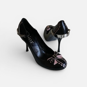 以下が含まれることがあります： 黒いハイヒールパンプス。リボンとヒールに「Dior」の文字がデザインされています。パンプスはポインテッドトゥで、細いピンヒールです。リボンはピンク色で白い水玉模様。サイドにはシルバーチェーンのディテールがあります。
