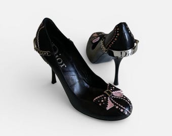 Könnte beinhalten: Schwarze High-Heels-Pumps mit Schleifendetail und dem Wort "Dior" auf der Ferse. Die Pumps haben eine spitze Zehenpartie und einen dünnen Stiletto-Absatz. Die Schleife ist rosa mit weißen Punkten. Ein silbernes Kettendetail befindet sich an der Seite.