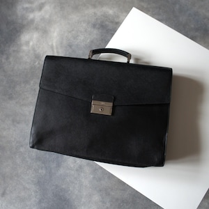 May include: A black leather briefcase with a textured surface, featuring a single top handle and a rectangular flap closure. The front is secured with a silver-toned metal clasp that includes a central keyhole. The design is structured and rectangular.