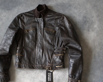 Könnte beinhalten: Braune Lederjacke mit Stehkragen, mehreren Reißverschlusstaschen und Schnallendetails. Die Jacke hat ein kurzes Design und ein Etikett mit der Aufschrift "VERSACE JEANS COUTURE".