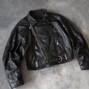 May include: Black leather jacket with an asymmetrical zip, multiple zipped pockets, and a belted waist. The jacket has silver-tone hardware and decorative perforations on the sleeves. A classic biker-style jacket.