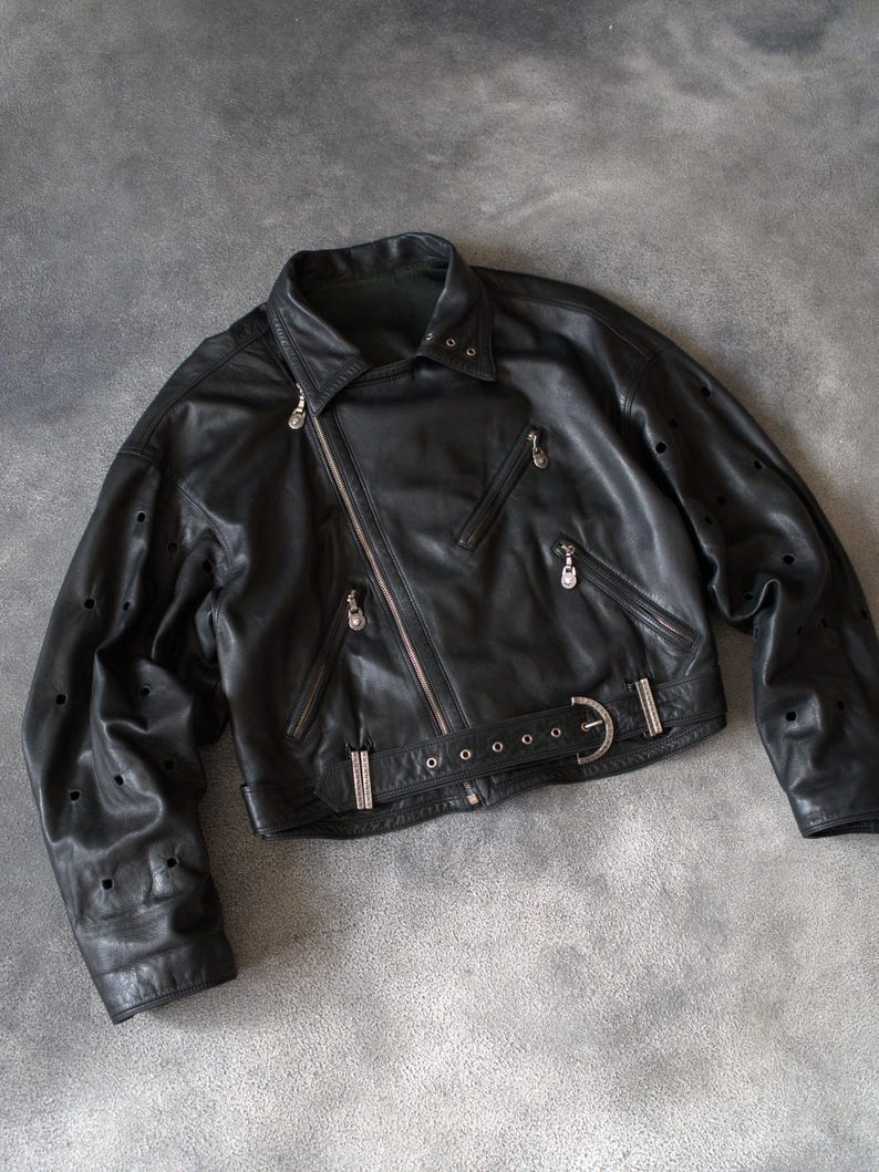May include: Black leather jacket with an asymmetrical zip, multiple zipped pockets, and a belted waist. The jacket has silver-tone hardware and decorative perforations on the sleeves. A classic biker-style jacket.