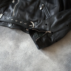 May include: A black leather jacket with silver zips and a belt. The jacket has a classic biker style with a notched collar, multiple pockets, and perforated details on the sleeves. The jacket is shown on a grey surface.