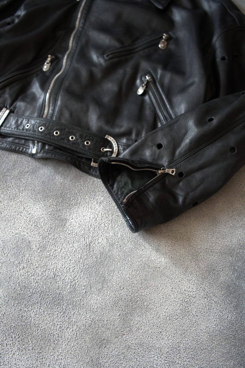 May include: A black leather jacket with silver zips and a belt. The jacket has a classic biker style with a notched collar, multiple pockets, and perforated details on the sleeves. The jacket is shown on a grey surface.