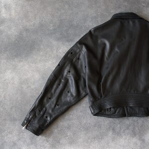 May include: Black leather jacket with a classic collar and a ribbed hem. The jacket features a zip closure on the sleeve and decorative perforations along the sleeve. A stylish outerwear piece.