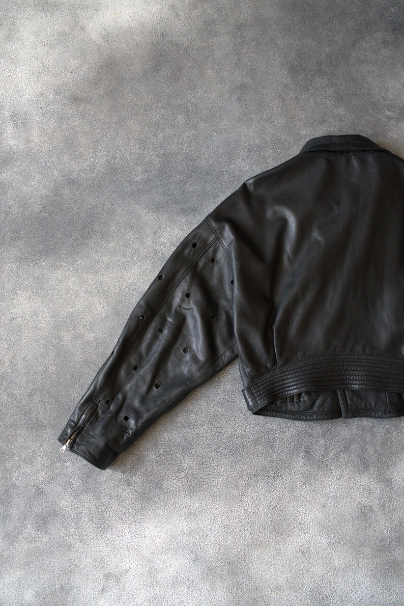 May include: Black leather jacket with a classic collar and a ribbed hem. The jacket features a zip closure on the sleeve and decorative perforations along the sleeve. A stylish outerwear piece.