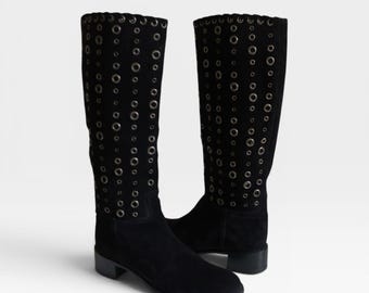 Könnte beinhalten: Schwarze Kniehohe Stiefel aus Wildleder. Die Stiefel haben silberfarbene Ösen entlang des Schafts und einen kleinen Blockabsatz. Die Stiefel haben eine runde Spitze und ein mattes Finish. Ungefähre Höhe: 40 cm.