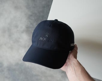 Könnte beinhalten: Marineblaue Baseballkappe mit dem gestickten Wort "ROLEX" und einem Kronenlogo auf der Vorderseite. Das Wort "SAL" ist auf der Seite gestickt. Die Kappe ist aus glattem Stoff gefertigt.