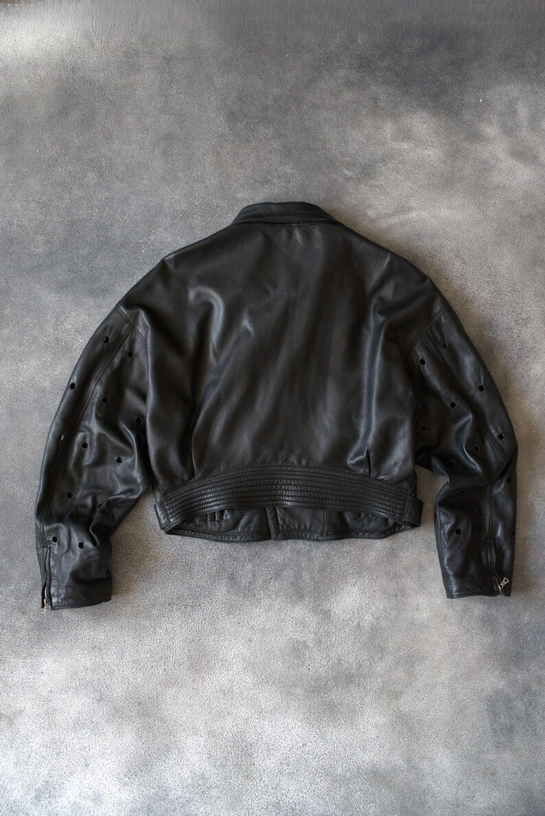 May include: A black leather jacket, cropped in style. It has a ribbed waistband and cuffs, a classic collar, and perforated detailing on the sleeves. The rear view highlights the jacket's design and construction.