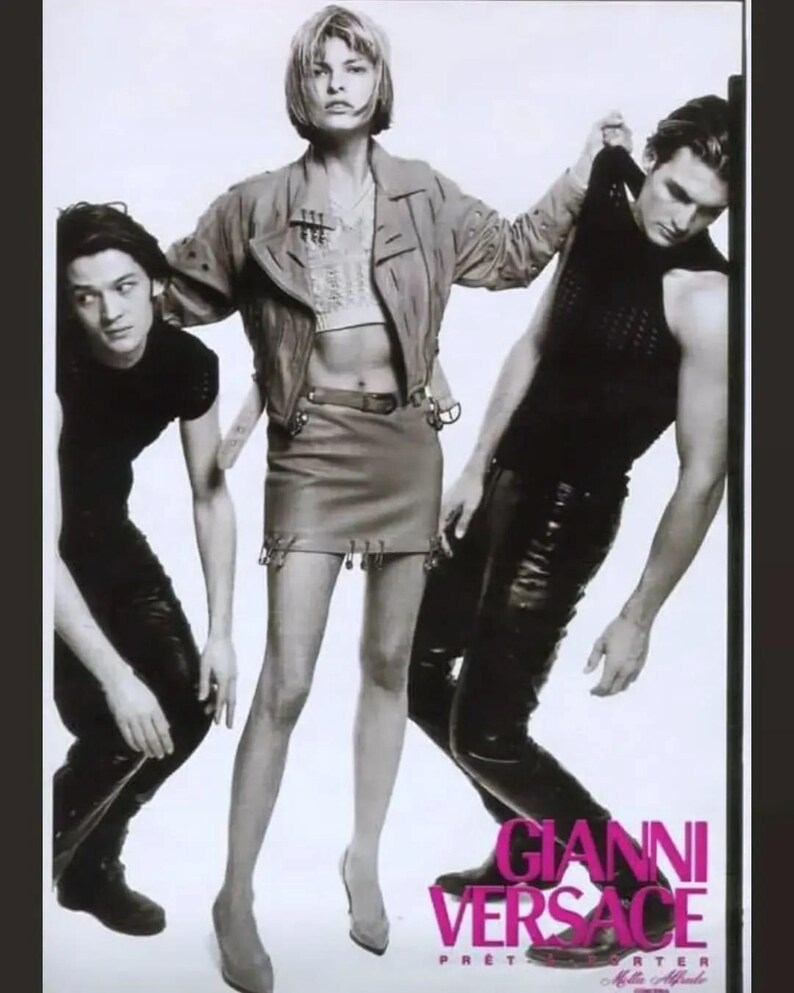 May include: Black and white fashion advertisement featuring three people. A woman in a cropped jacket and skirt stands between two people in black clothing. The text "GIANNI VERSACE" is displayed in pink and white.