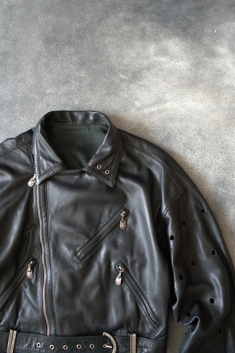 May include: Black leather jacket with an asymmetrical zip, multiple zipped pockets, and a notched collar. The jacket has a belt at the waist and decorative perforations on the sleeves.