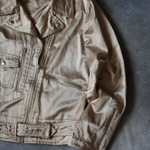 Op de afbeelding: Beige satijnen bikerjack met een glanzende afwerking. Het heeft een reverskraag, meerdere zakken met ritssluiting en een ceintuur in de taille. De jas heeft een subtiele glans en de merknaam "miu miu" op het label.