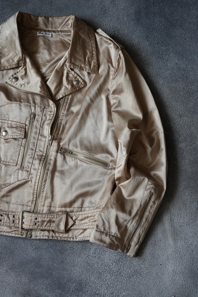 Op de afbeelding: Beige satijnen bikerjack met een glanzende afwerking. Het heeft een reverskraag, meerdere zakken met ritssluiting en een ceintuur in de taille. De jas heeft een subtiele glans en de merknaam "miu miu" op het label.