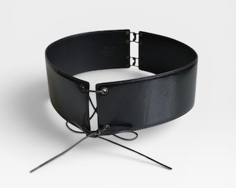 Könnte beinhalten: Breiter, schwarzer Gürtel mit glänzendem Finish. Der Gürtel hat ein Schnürdesign vorne und einen Schnallenverschluss hinten. Der Gürtel ist so konzipiert, dass er die Taille zusammenzieht und jedem Outfit einen stilvollen Akzent verleiht.