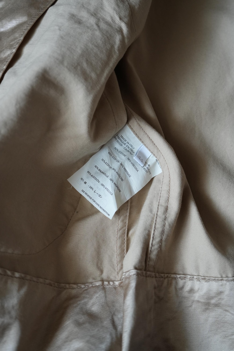 Op de afbeelding: Close-up van de binnenkant van een beige kledingstuk. Een wit label is zichtbaar, met daarop de samenstelling van het kledingstuk: 62% katoen, 38% viscose. Het label vermeldt ook "Made in Italy".