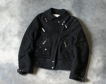 Könnte beinhalten: Schwarze Wildlederjacke mit silbernen Reißverschlüssen, Nieten und Knopfverschluss. Die Jacke hat einen Kragen, zwei Vordertaschen und verstellbare Schnallen an den Ärmeln. Ein stilvolles Oberbekleidungsstück.