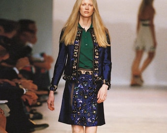 Könnte beinhalten: Ein Model auf einem Laufsteg, das eine marineblaue Jacke mit dekorativen Verzierungen, ein grünes Poloshirt und einen marineblauen Rock mit Paillettenbesatz trägt. Das Model hat lange blonde Haare und geht auf den Betrachter zu.