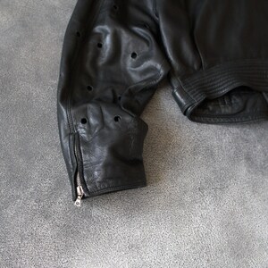 May include: Black leather jacket with perforated detailing on the sleeve. The jacket has a zip cuff and a ribbed hem. The leather appears soft and worn. A classic style jacket.