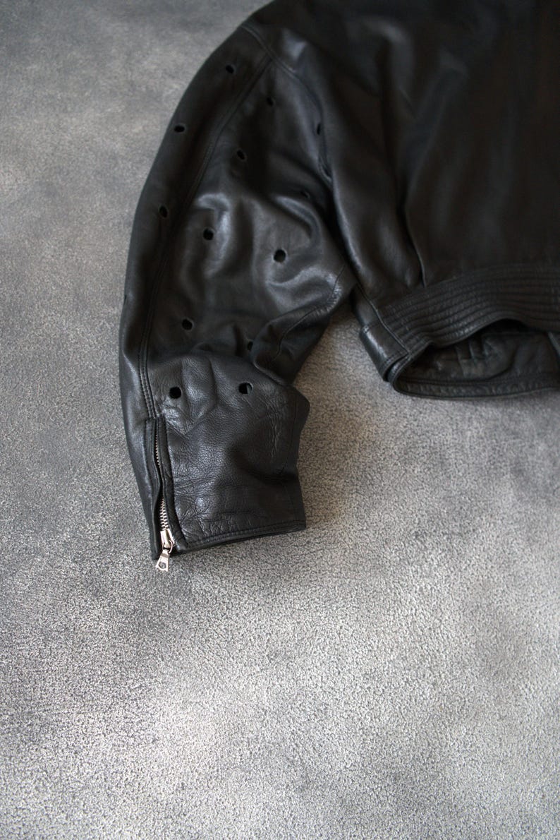 May include: Black leather jacket with perforated detailing on the sleeve. The jacket has a zip cuff and a ribbed hem. The leather appears soft and worn. A classic style jacket.