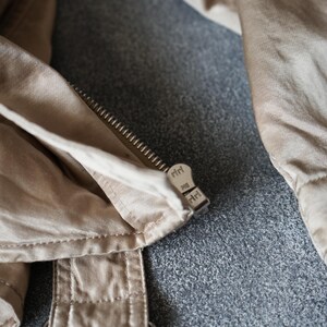 Op de afbeelding: Close-up van een beige jas met een metalen rits en gesp. De ritstrekker is gemarkeerd met de letters "riri". De jasstof is lichtbeige van kleur.