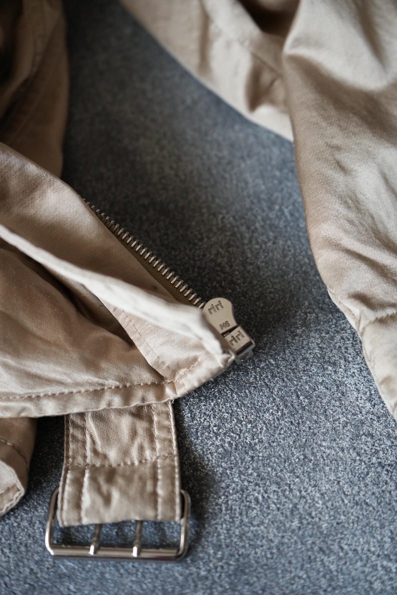Op de afbeelding: Close-up van een beige jas met een metalen rits en gesp. De ritstrekker is gemarkeerd met de letters "riri". De jasstof is lichtbeige van kleur.