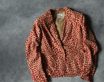 Könnte beinhalten: Gemusterter Blazer in Beige und Rot mit sich wiederholendem Blattdesign. Die Jacke hat ein fallendes Revers, einen Ein-Knopf-Verschluss und ein beiges Futter. Der Blazer liegt flach auf einer grauen Oberfläche. Ein stilvolles Kleidungsstück.