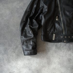 May include: Black leather jacket with a zip fastening and silver hardware. The jacket has a notched collar, a belt at the waist, and decorative perforations on the sleeves. The jacket is shown on a grey background.