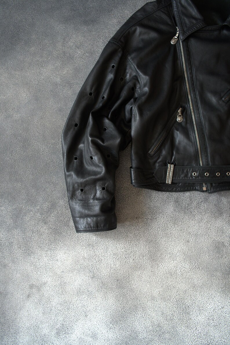 May include: Black leather jacket with a zip fastening and silver hardware. The jacket has a notched collar, a belt at the waist, and decorative perforations on the sleeves. The jacket is shown on a grey background.