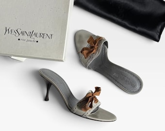 May include: A pair of light gray leather mules with brown ribbon bows, featuring perforated brogue detailing and thin heels. The shoes are accompanied by a white Yves Saint Laurent Rive Gauche branded box and a black satin dust bag.