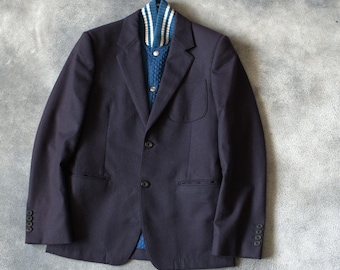 Könnte beinhalten: Ein marineblauer Blazer mit einem blauen Pullover darunter. Der Pullover hat ein Zopfmuster und einen weiß-blau gestreiften Kragen. Der Blazer hat drei Knöpfe an jeder Manschette und zwei Vordertaschen.