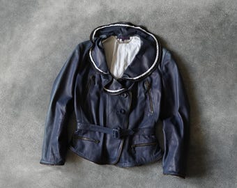 Könnte beinhalten: Eine dunkelblaue Lederjacke mit einem großen Rüschenkragen mit weißem Rand, einer Knopfleiste und einem passenden Taillengürtel mit Schnalle. Die Jacke verfügt über zwei Reißverschlusstaschen auf der Brust und zwei an der Taille sowie ein weiß gestreiftes Innenfutter.