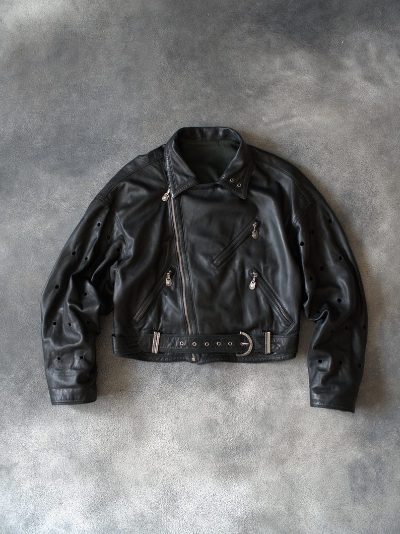 May include: Black leather biker jacket with an asymmetrical zip, notched collar, and waist belt. The jacket has decorative perforations on the sleeves and silver-toned metal hardware. A classic, stylish outerwear piece.
