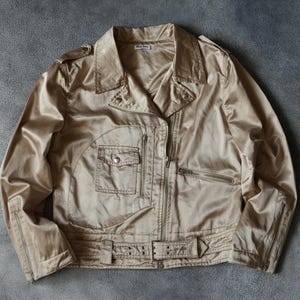 Op de afbeelding: Een beige jas met een glanzende, satijnachtige afwerking. De jas heeft een klassieke bikerstijl met een reverskraag, een ritssluiting en meerdere zakken. De jas heeft een riem in de taille en ritsen op de mouwen.