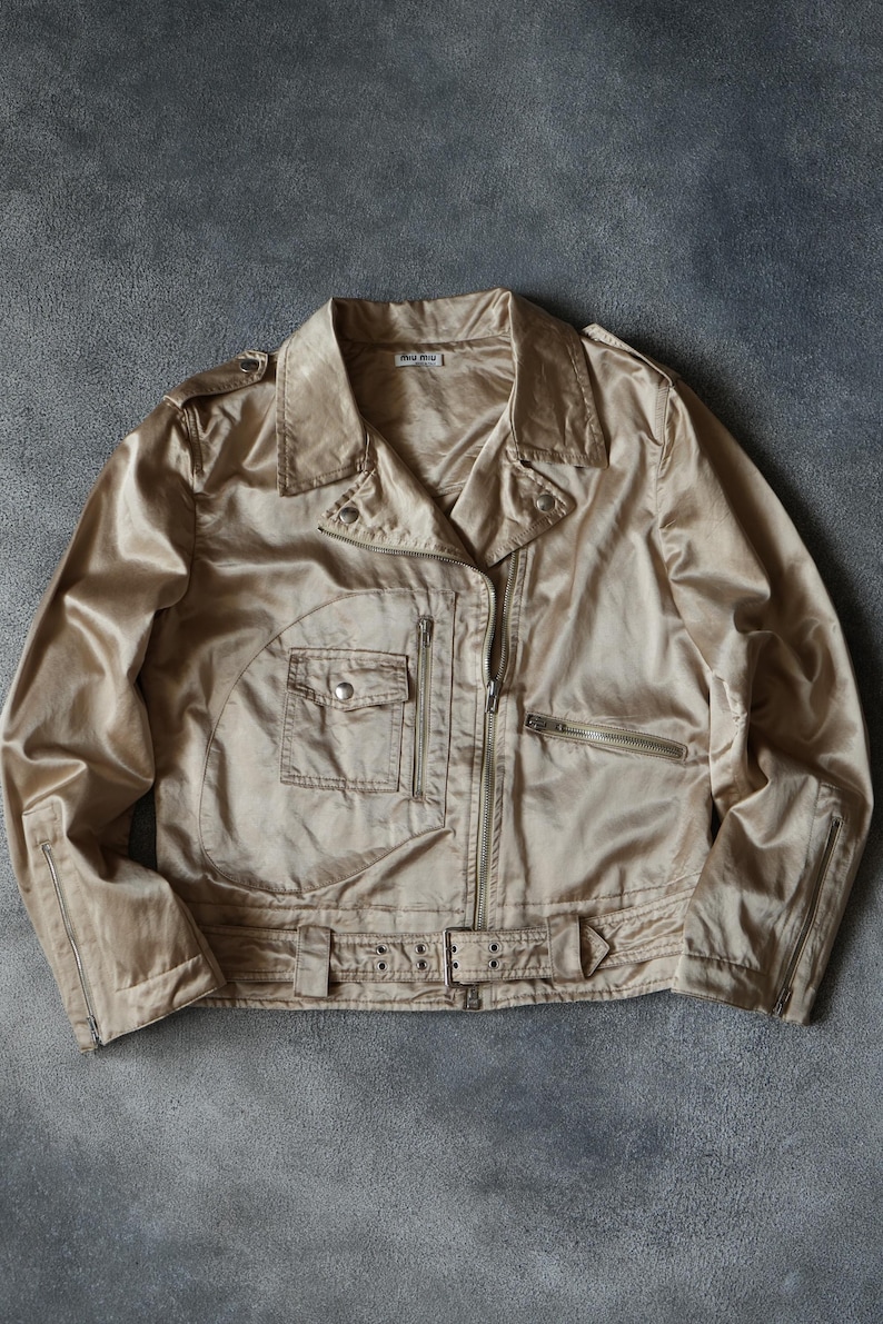 Op de afbeelding: Een beige jas met een glanzende, satijnachtige afwerking. De jas heeft een klassieke bikerstijl met een reverskraag, een ritssluiting en meerdere zakken. De jas heeft een riem in de taille en ritsen op de mouwen.