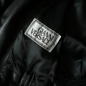 May include: Close-up of a black leather jacket interior. A white label with the text "GIANNI VERSACE" in black lettering is sewn into the lining. The jacket's interior is a smooth, dark fabric.