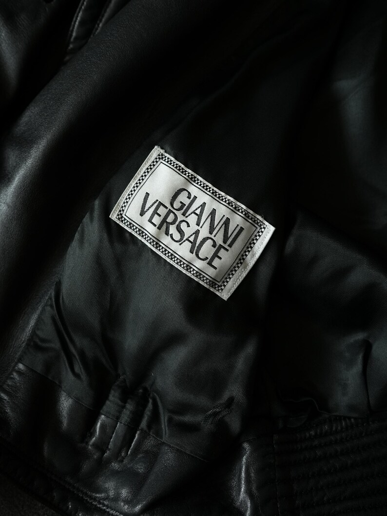 May include: Close-up of a black leather jacket interior. A white label with the text "GIANNI VERSACE" in black lettering is sewn into the lining. The jacket's interior is a smooth, dark fabric.