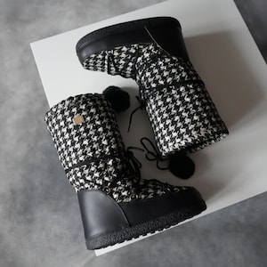 Pode incluir: Um par de botas de inverno com estampa pied-de-poule em preto e branco. As botas têm solas e detalhes em preto, com design de cadarço e pequenos pompons pretos. As botas são feitas de material acolchoado.