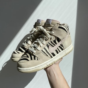 Puede incluir: Zapatillas altas beige y negras con estampado de cebra. Las zapatillas tienen una suela color crema, cordones y un logotipo de swoosh rosa. Los zapatos son de ante y cuero.