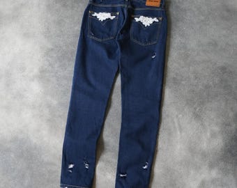 Könnte beinhalten: Dunkelblaue Jeans mit weißer Blumenstickerei auf den Gesäßtaschen. Die Jeans hat Distressed-Details an Knien und Knöcheln. Ein braunes Etikett ist hinten am Bund sichtbar.
