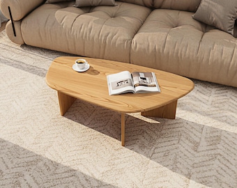 May include: A light-colored wooden coffee table with a rounded shape, featuring a book and a cup of coffee on top. The table is set on a patterned rug, with a beige sofa in the background.