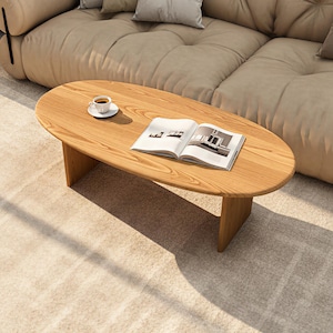 Puede incluir: Mesa de centro ovalada de madera clara. Una pequeña taza y platillo blancos con café están sobre la mesa, junto con una revista abierta. La mesa está frente a un sofá seccional beige.
