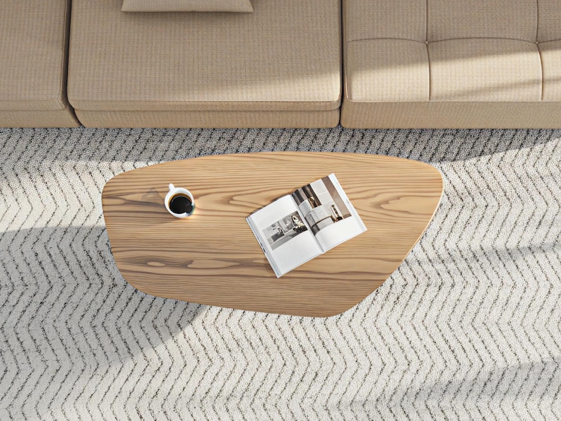 May include: Overhead view of a light wood coffee table with a cup of coffee and an open magazine. The table sits on a patterned rug, near a beige sofa. The coffee table has a unique, organic shape.