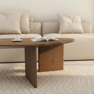 May include: A modern living room scene featuring a wooden coffee table with a rounded edge and a solid base. A small white teacup and saucer sit on the table, along with an open book. The table is in front of a beige sofa with matching pillows.