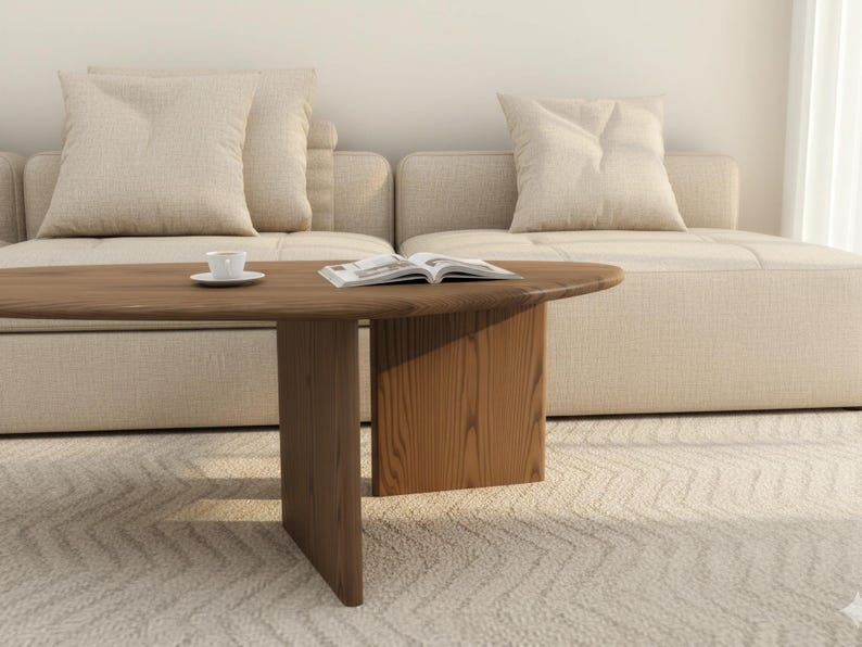 May include: A modern living room scene featuring a wooden coffee table with a rounded edge and a solid base. A small white teacup and saucer sit on the table, along with an open book. The table is in front of a beige sofa with matching pillows.