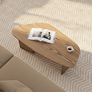 May include: A wooden coffee table with an open book and a small white coffee cup. The table is light brown and has an unusual shape. It sits on a patterned rug. A section of a beige sofa is visible.