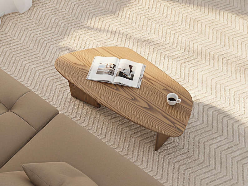 May include: A light brown wooden coffee table with an open book and a small white coffee cup. The table has a unique shape and is placed on a patterned rug. A section of a beige sofa is visible.