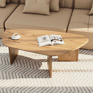 May include: A light-colored wooden coffee table with an oval shape and three legs. An open book and a cup of coffee on a saucer are on the table. The table is in front of a beige sofa.