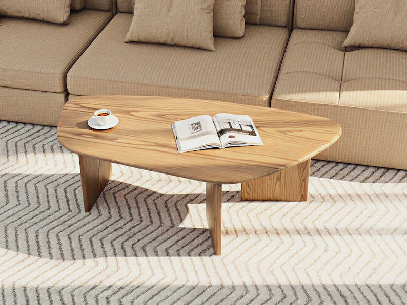 May include: A light-colored wooden coffee table with an oval shape and three legs. An open book and a cup of coffee on a saucer are on the table. The table is in front of a beige sofa.