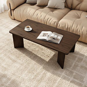 May include: A dark brown wooden coffee table with a rectangular top and block legs. A white coffee cup and saucer sit on the table, along with an open magazine. The table is placed on a cream-colored rug, near a beige sofa.