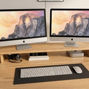 May include: Two silver iMac computers displaying a mountain landscape are on a light wood desk. A wooden monitor stand holds the computers. A keyboard, mouse, and black mousemat are in front. A camera and books are on the desk.